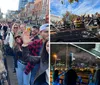 A lively group of people is smiling and posing for a photo with drinks in their hands on a bustling city street lined with bars and shops