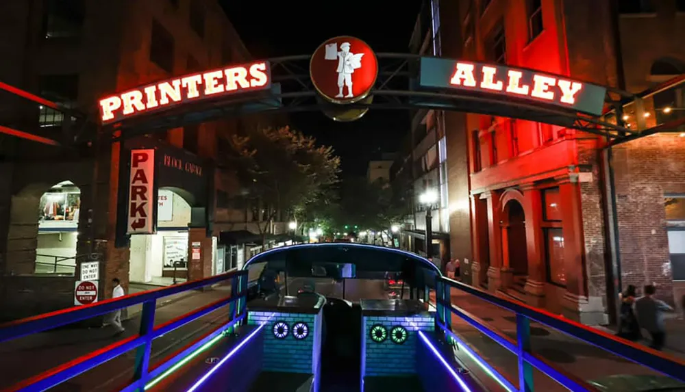 The image shows a neon-lit archway marking the entrance to Printers Alley at night with the interior of a vehicle in the foreground featuring illuminated blue lights