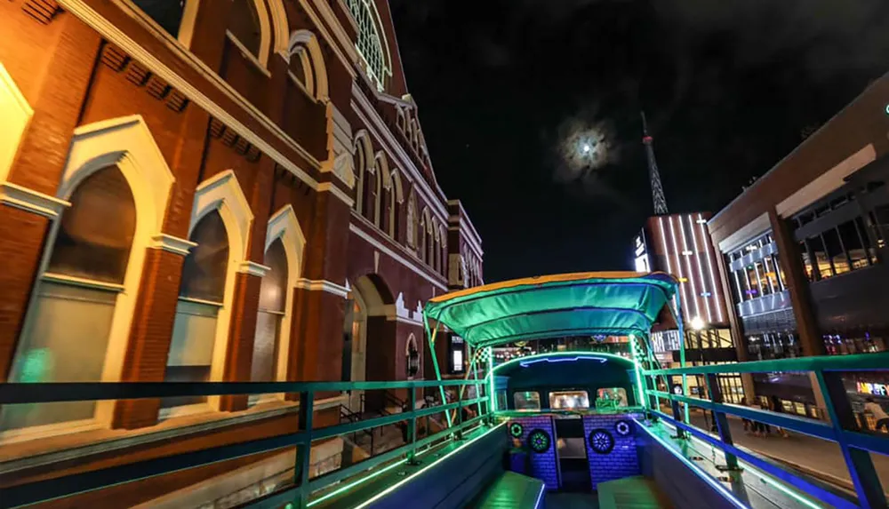 A colorful open-top bus is parked on a street at night with illuminated buildings in the background and a cloudy night sky above