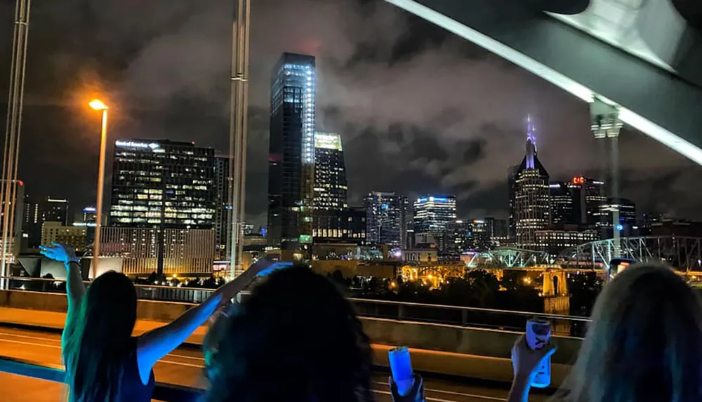 Silhouetted figures are raising their hands against the backdrop of a brightly lit cityscape at night under a cloudy sky