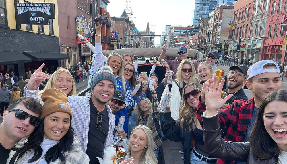 A lively group of people is smiling and posing for a photo with drinks in their hands on a bustling city street lined with bars and shops