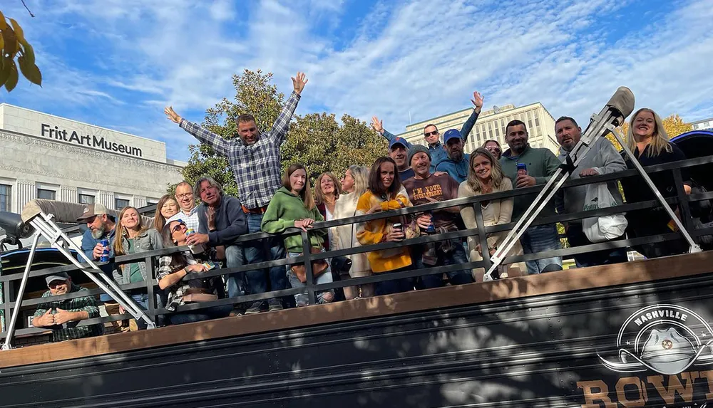 A group of cheerful people is standing on a double-decker tour bus waving and enjoying themselves in front of the Frist Art Museum