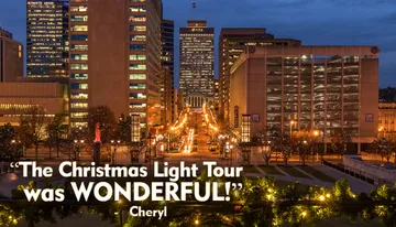 The image captures a dusk view of a city street lined with illuminated buildings, decorative lights, and a statue, all framed by festive garland in the foreground.