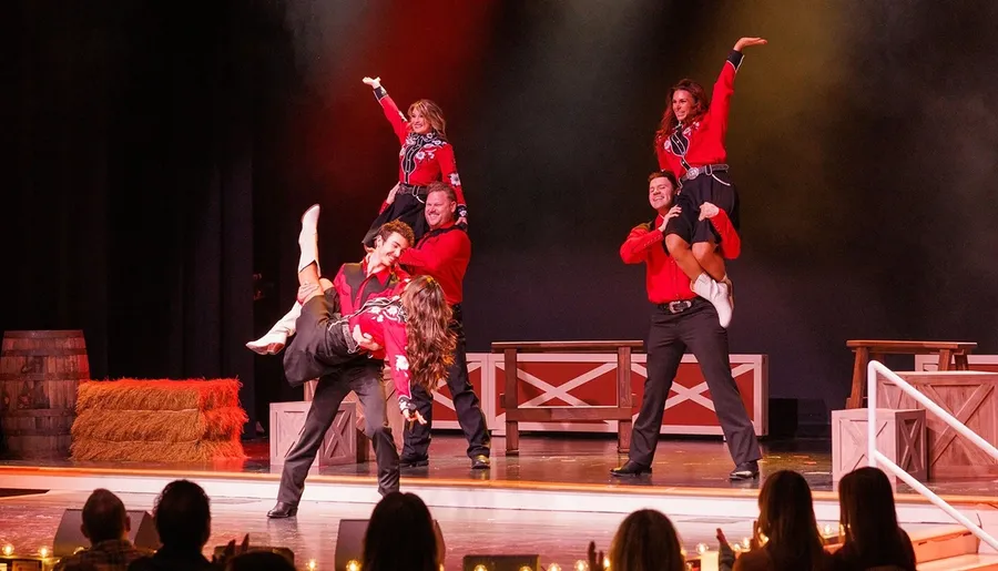 Five performers in matching red and black outfits engage in an energetic dance routine on stage, with some dancers lifted in the air, in front of an audience in a theater setting.