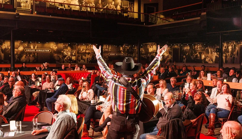 A musician in a colorful shirt and cowboy hat stands on a stage with a guitar facing a cheering audience in a lively venue