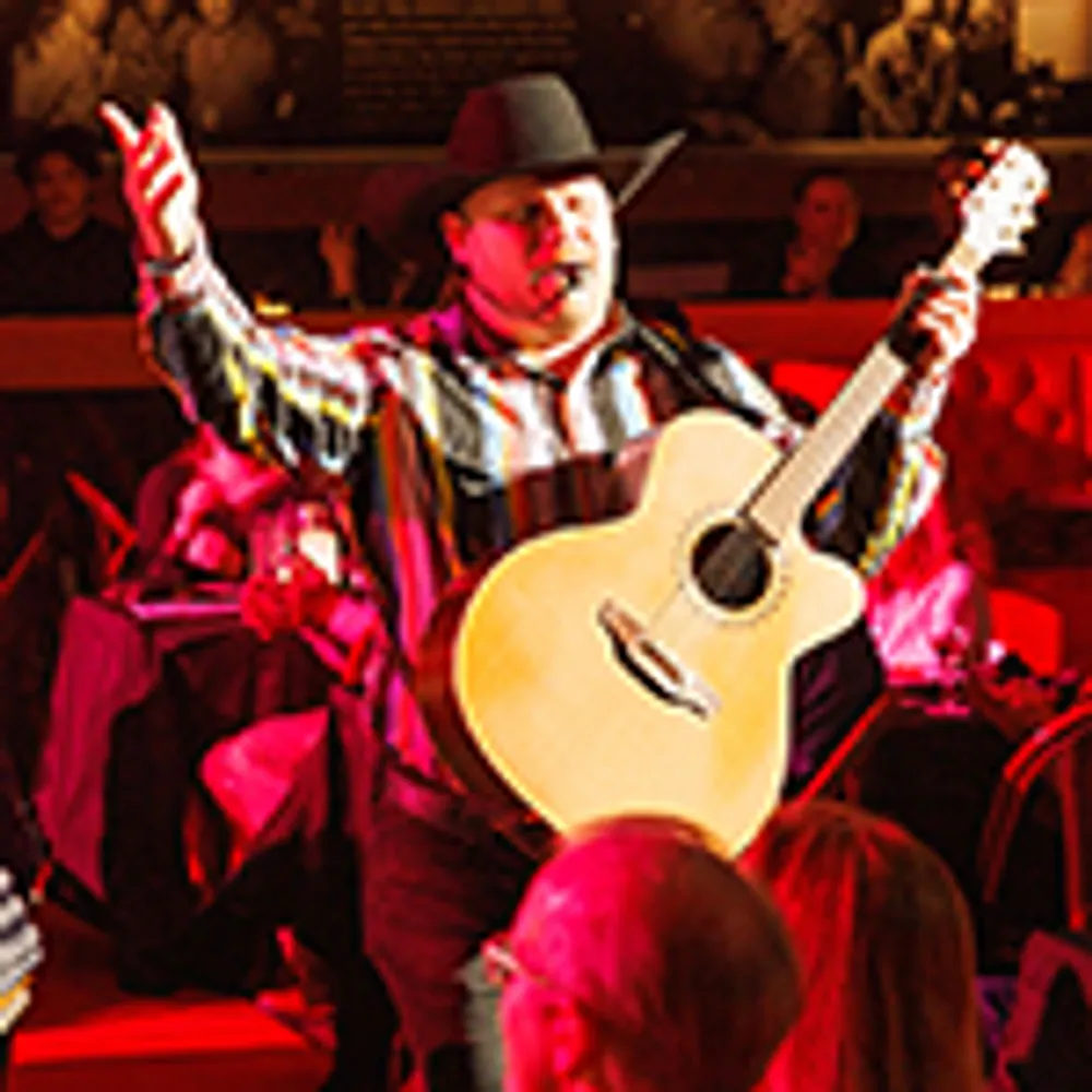 A performer in a cowboy hat is playing a guitar and singing to an audience in a vibrant red-lit setting