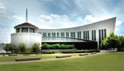 The image shows the Country Music Hall of Fame and Museum, with its modern architectural design and distinctive windows, located in a large open space with grass and trees under a partly cloudy sky.