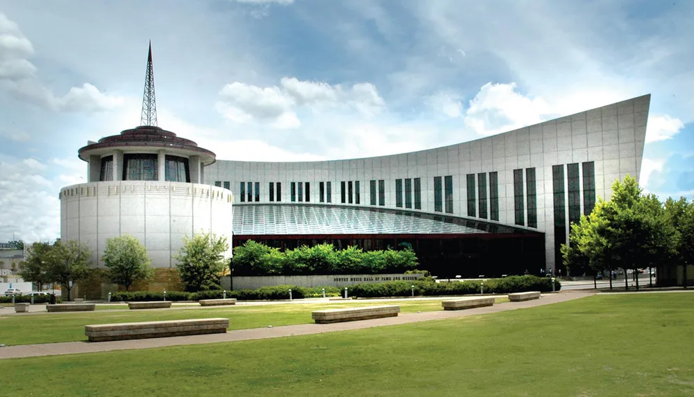The image shows the Country Music Hall of Fame and Museum with its modern architectural design and distinctive windows located in a large open space with grass and trees under a partly cloudy sky