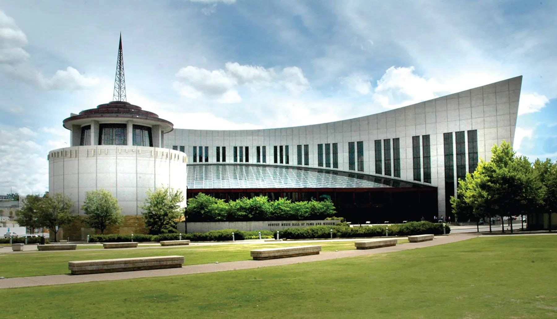 The image shows the Country Music Hall of Fame and Museum, with its modern architectural design and distinctive windows, located in a large open space with grass and trees under a partly cloudy sky.