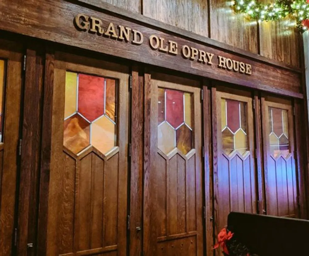 The image shows the richly colored wooden entrance doors to the Grand Ole Opry House accented with stained glass and adorned with a sign of the venues name overhead