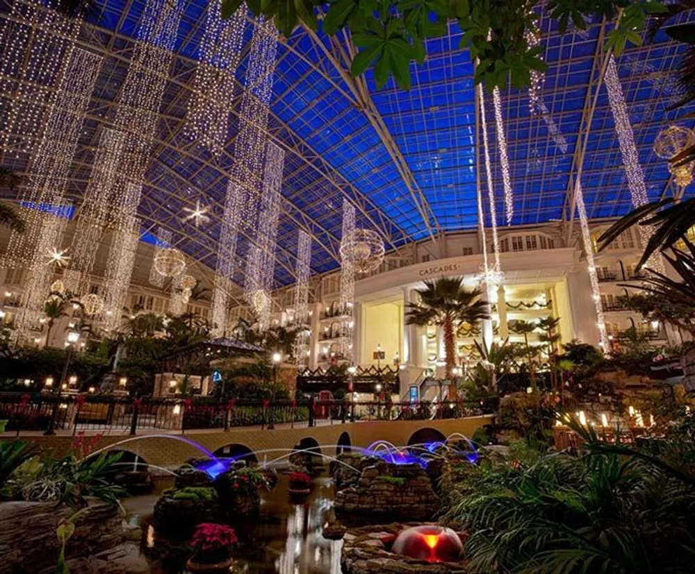 The image shows an indoor space with a glass ceiling adorned with cascading lights and surrounded by lush greenery and a small water feature creating a tranquil festive atmosphere
