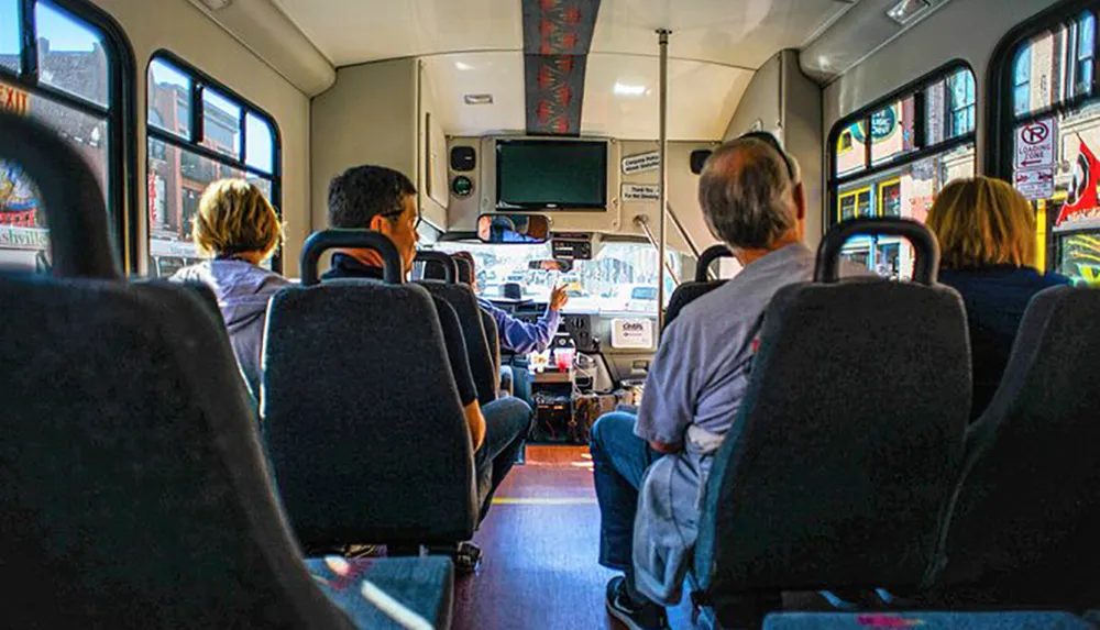Passengers are seated in a bus facing forward towards the driver who is gesturing with his hand with a view of the street ahead visible through the windshield