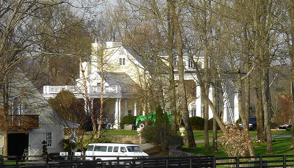 The image shows a large two-story house partly obscured by trees with a white passenger van parked in the driveway and a large green tractor visible in the yard