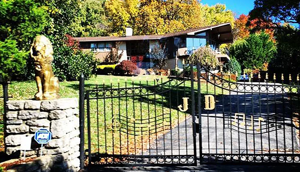 An ornate gated entrance with the initials JD leads to a residential property with a mid-century modern house and a statue on the front lawn