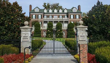 The image shows a grand, brick estate with a gated entrance, surrounded by trimmed hedges and trees.