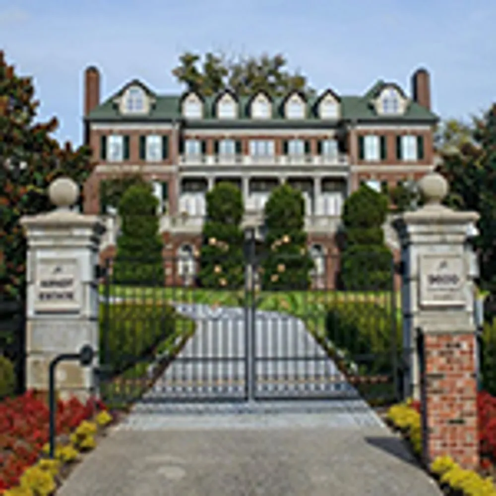 The image shows a large elegant brick mansion with a gated entrance and well-manicured landscaping