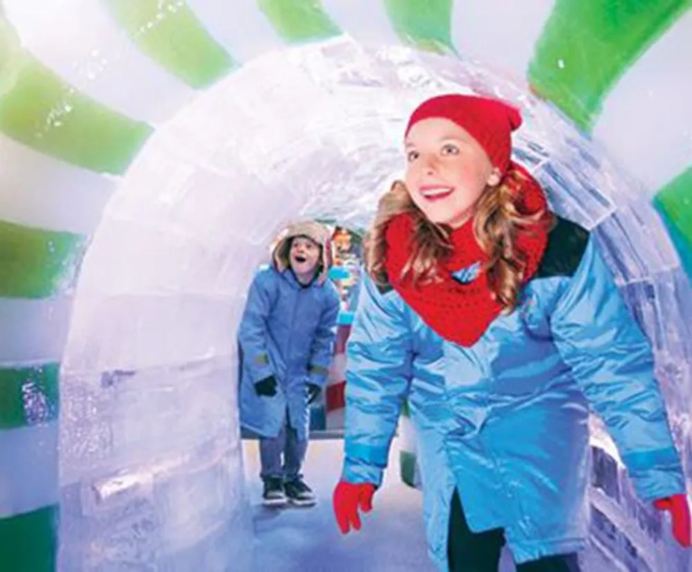 Two children in winter attire are enjoying themselves as they explore a colorful ice tunnel