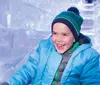 A child is joyfully sliding down a clear ice slide wearing winter clothes and a big smile