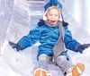 A child is joyfully sliding down a clear ice slide wearing winter clothes and a big smile