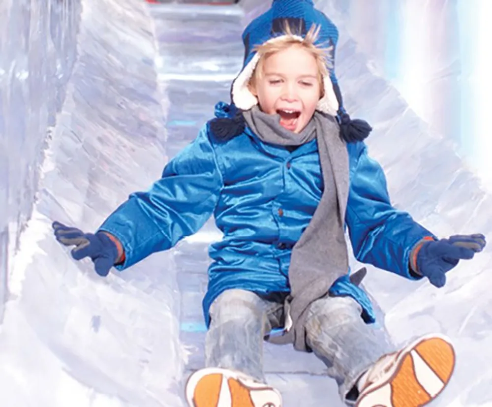 A child is joyfully sliding down a clear ice slide wearing winter clothes and a big smile