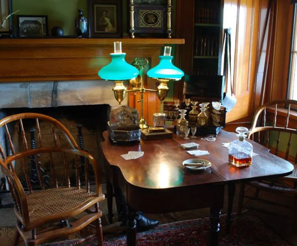 The image depicts a vintage room set up with a wooden table two chairs a couple of green-shaded lamps playing cards and decanters suggesting a setting for a period-style leisure activity or game
