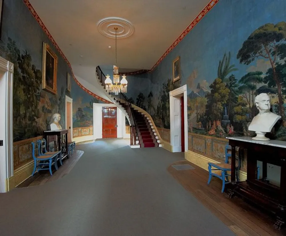 The image shows a grand room with an elegant curved staircase decorative wall murals a chandelier sculptures and antique furniture conveying a sense of historical opulence