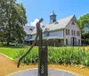 An old-fashioned water pump sits in the foreground with a charming large two-story house with a pointed roof and turret in the background surrounded by greenery under a blue sky