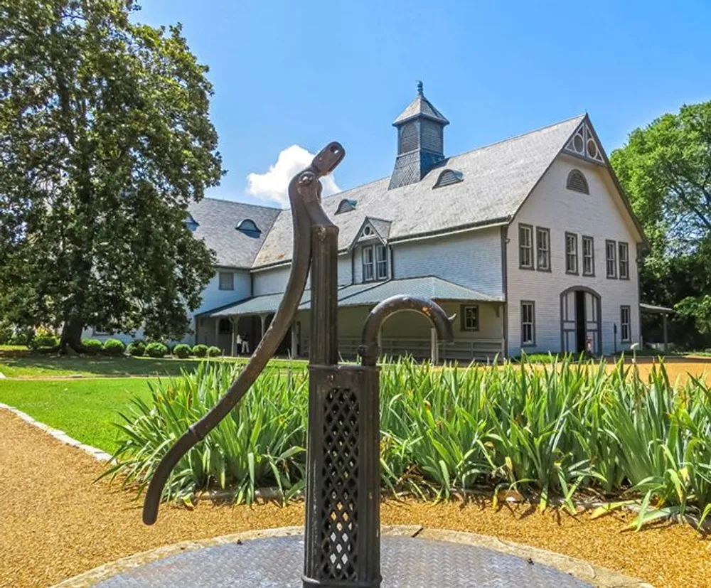 An old-fashioned water pump sits in the foreground with a charming large two-story house with a pointed roof and turret in the background surrounded by greenery under a blue sky