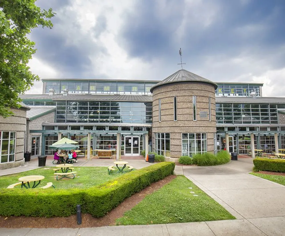 The image shows a modern building with a glass facade and a sign that says MARKET featuring a landscaped area with seating and a canopy-covered area where a few people are gathered