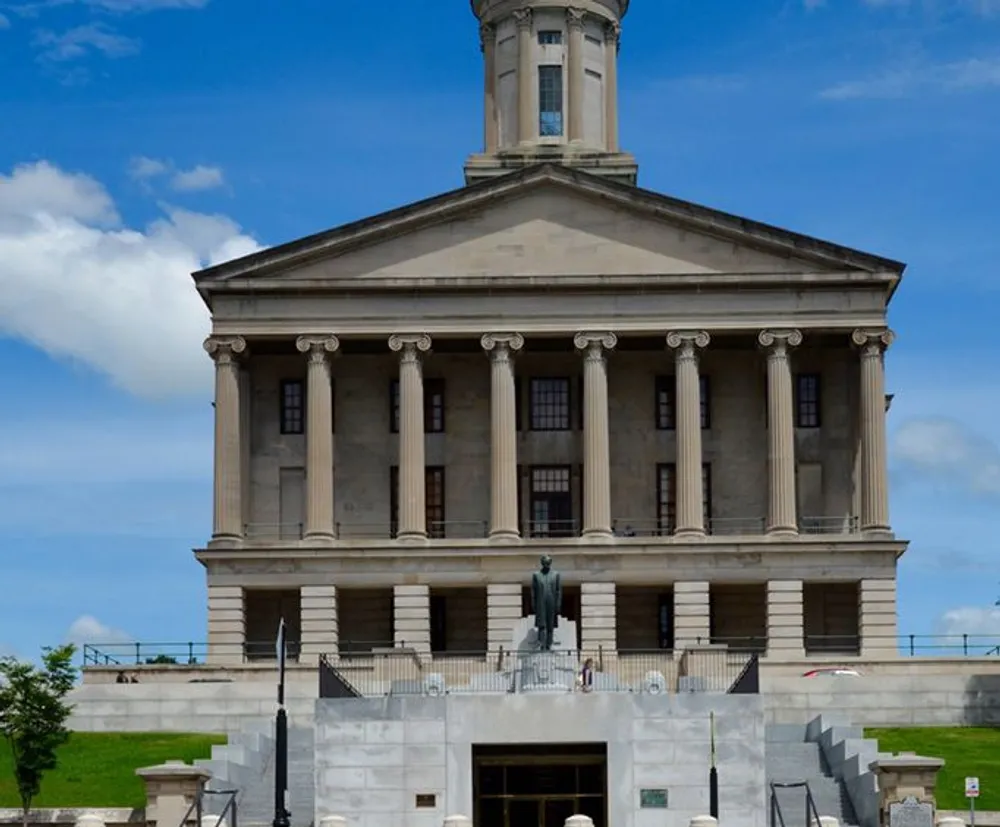 The image shows a stately classical government building with columns and a dome under a partly cloudy sky