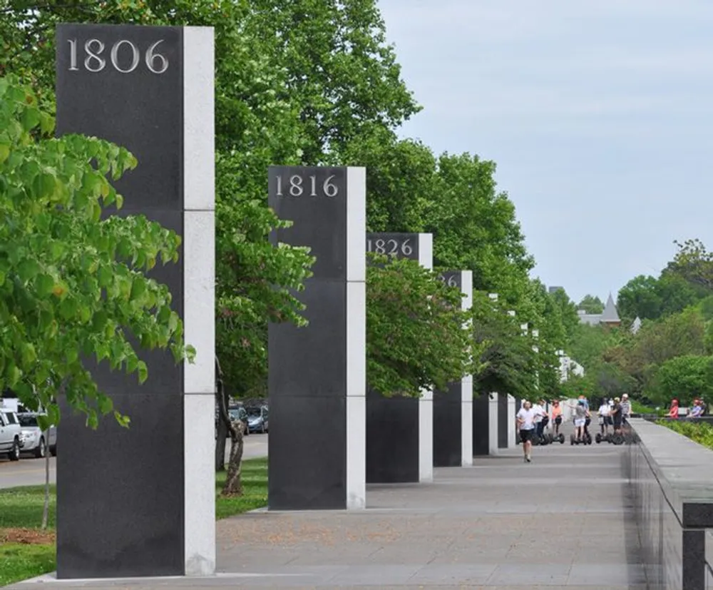 The image shows a row of pillars with different years marked on them aligned along a walkway with people and green trees in the background