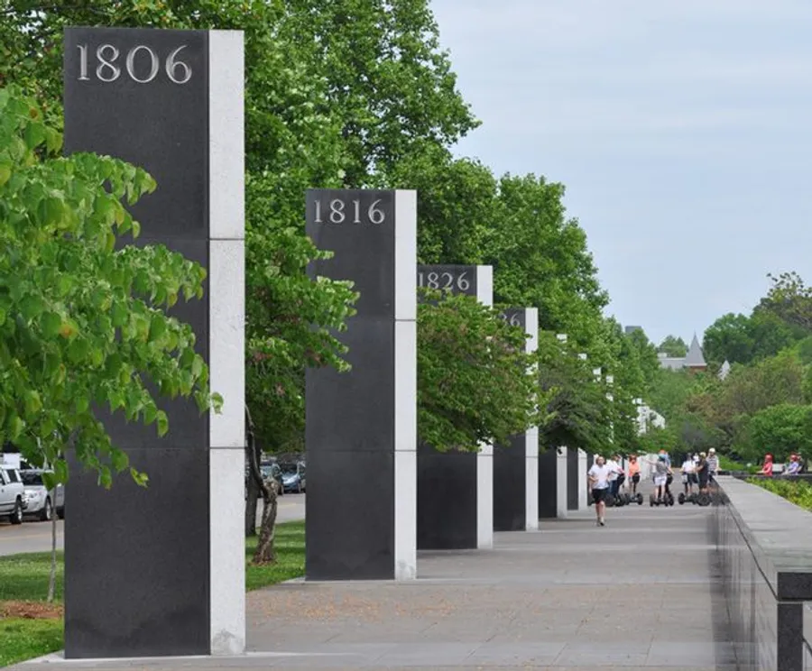 The image shows a row of pillars with different years marked on them, aligned along a walkway with people and green trees in the background.