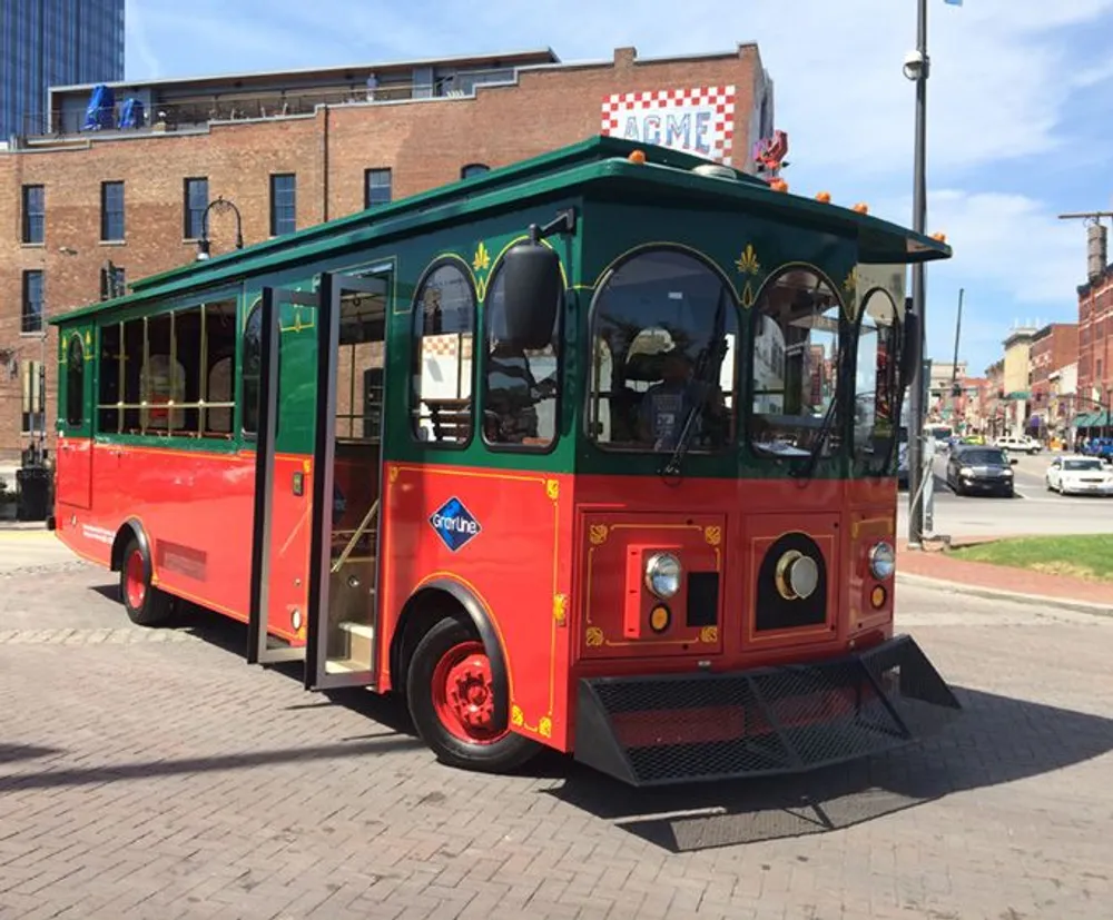 A red and green trolley-like tourist bus is parked on a sunny street with buildings in the background