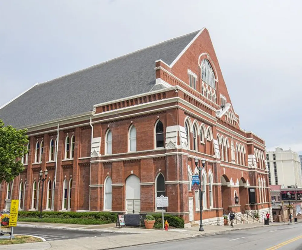 The image shows a large red brick Gothic Revival style building with arched windows and a steeply pitched roof situated at a street corner with overcast skies