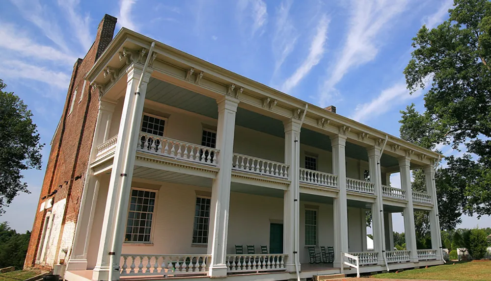 The image depicts a two-story colonial-style house with white columns and a large balcony set against a blue sky streaked with wispy clouds