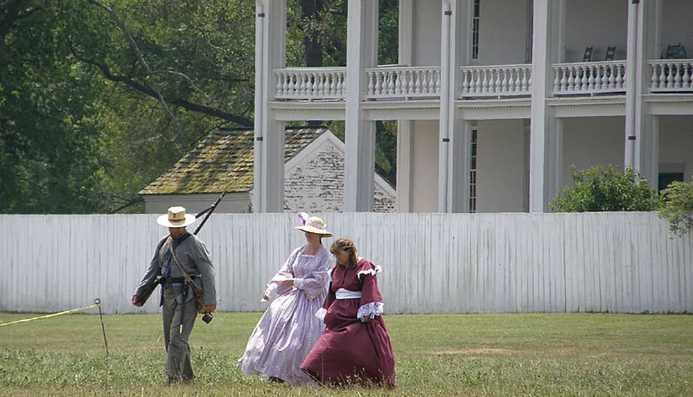 Two people dressed in historical clothing are walking across a grassy field near a white picket fence with a large white house in the background while a person in a soldiers uniform walks separately holding a rifle with a bayonet