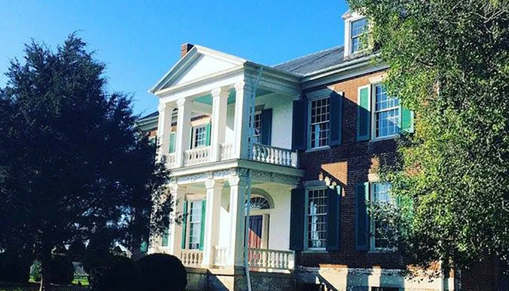 The image shows a two-story brick house with white columns and a balcony surrounded by trees under a clear blue sky