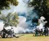 The image depicts a historical reenactment featuring individuals dressed in period military uniforms operating cannon artillery amidst billowing smoke likely simulating a battle scene