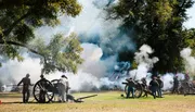 The image depicts a historical reenactment featuring individuals dressed in period military uniforms operating cannon artillery amidst billowing smoke, likely simulating a battle scene.