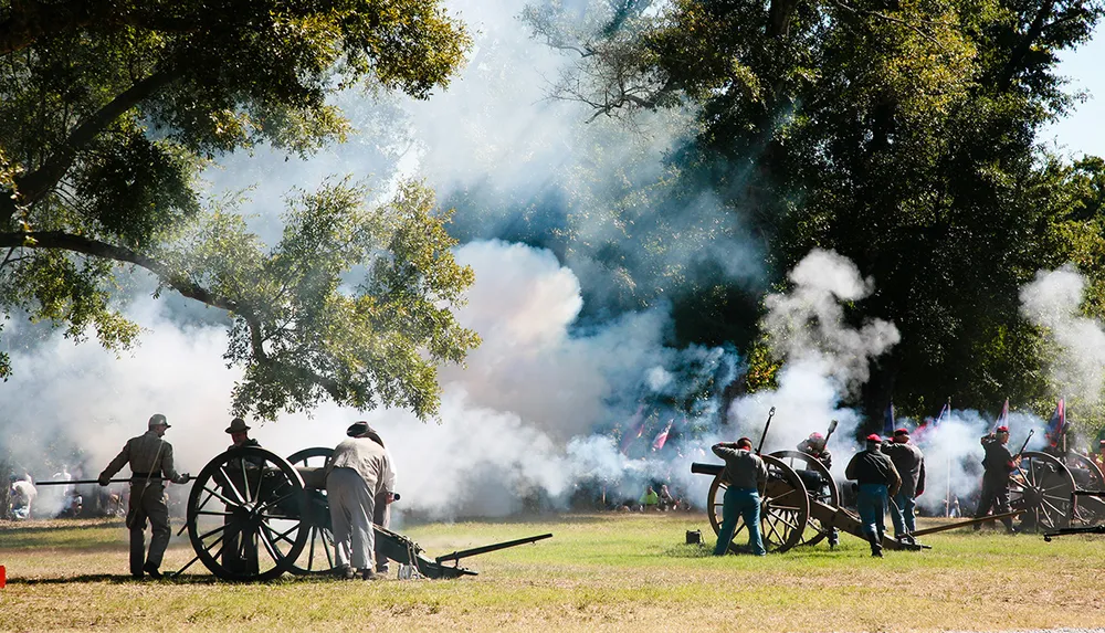 The image depicts a historical reenactment featuring individuals dressed in period military uniforms operating cannon artillery amidst billowing smoke likely simulating a battle scene