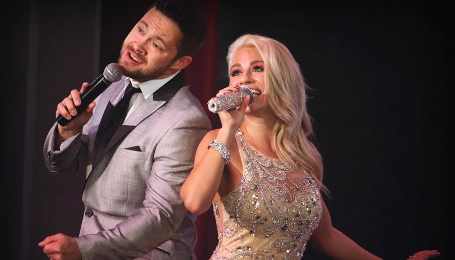 A man and a woman are performing on stage with microphones, likely singing or speaking, both dressed elegantly, with the woman in a sparkling dress.