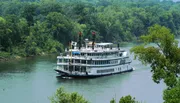 A paddle steamer is cruising along a river flanked by lush greenery.