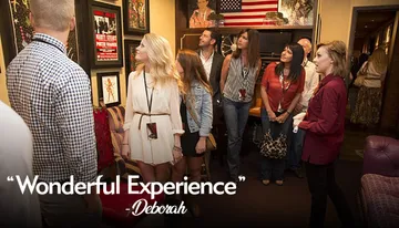 A group of people, seemingly on a guided tour, are attentively looking at framed posters and memorabilia on the walls of a room decorated with an American flag and various furnishings.