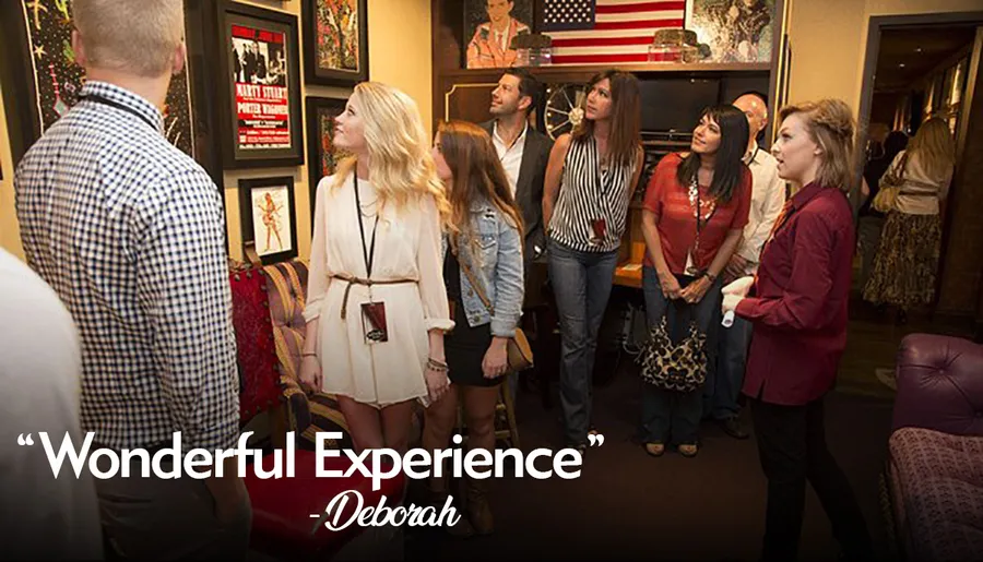 A group of people, seemingly on a guided tour, are attentively looking at framed posters and memorabilia on the walls of a room decorated with an American flag and various furnishings.