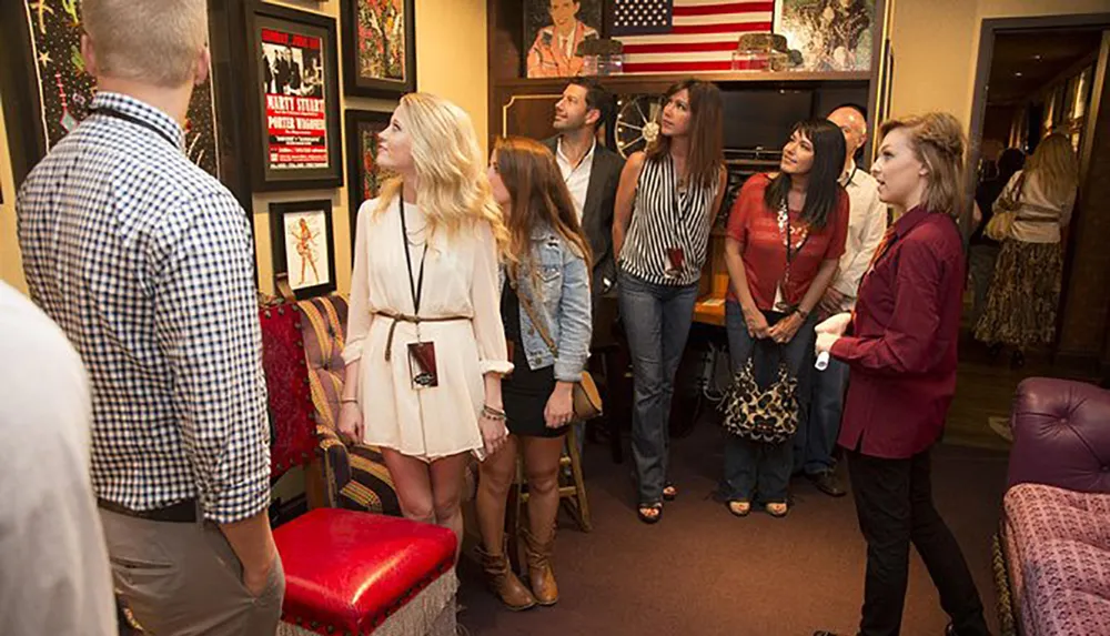 A group of people seemingly on a guided tour are attentively looking at framed posters and memorabilia on the walls of a room decorated with an American flag and various furnishings