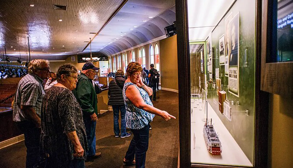 Visitors are observing exhibits in a museum gallery with one person pointing at a display case featuring a model ship