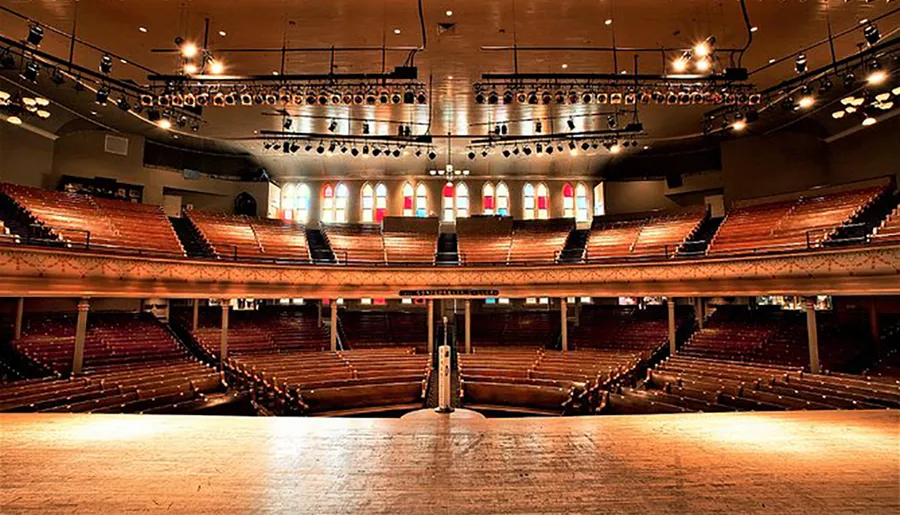 The image shows an empty grand theater with multiple tiers of seating, a large stage in the foreground, and stage lights overhead.