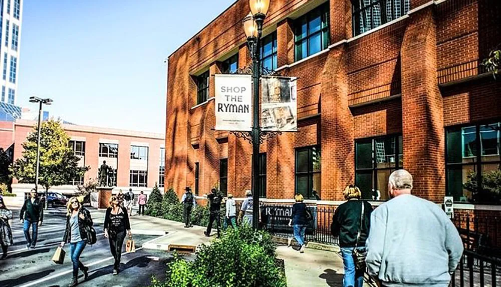 People are walking near a brick building that has a sign reading SHOP THE RYMAN suggesting the presence of a gift shop for the associated venue