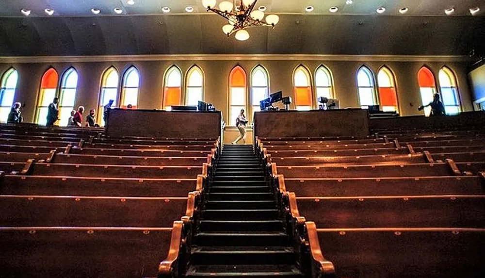 The image shows the interior of a church filled with rows of empty pews colorful stained-glass windows along the side and a few people scattered in the distance near the front