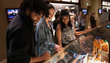 A group of people is smiling and looking at exhibits, possibly within a museum or gallery, where a guitar is displayed under glass.